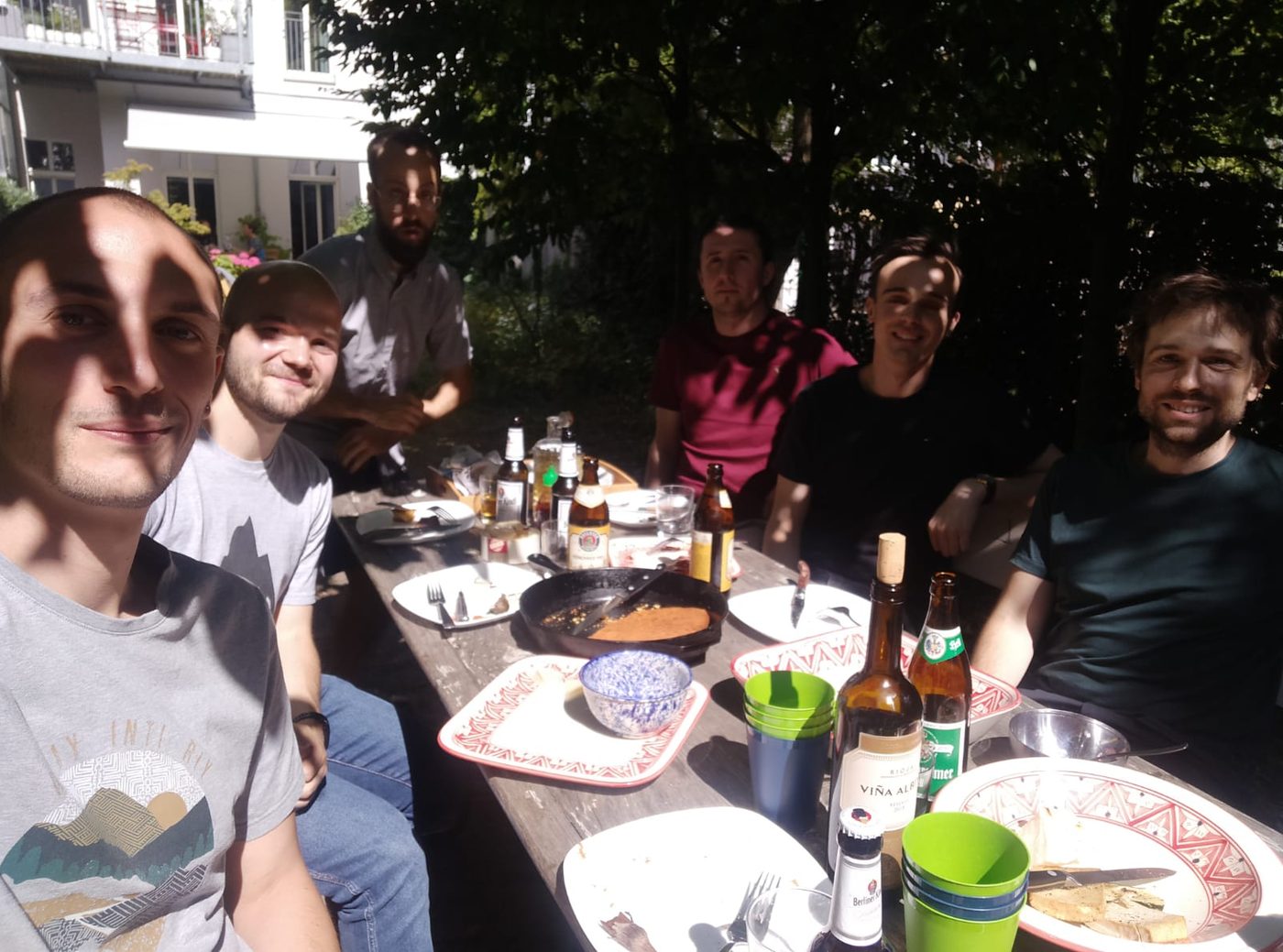 Six people around a garden table with the remains of a barbecue, in dappled summer shade.