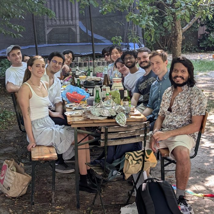 The group seated around a long table in a garden, under trees, at a summer barbecue.