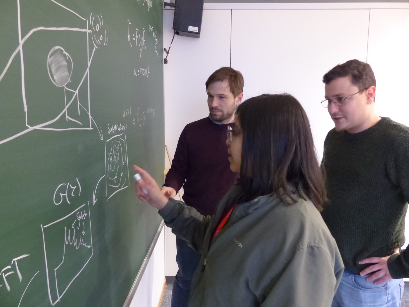 Three people at a blackboard covered in wave-optics lensing sketches and equations; one points at a drawing of lens substructure.